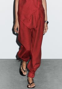 Pantaloni in lino rossi con una vestibilità ampia, con un dettaglio a nodo in vita. Abbinati a sandali neri. Sfondo grigio neutro.