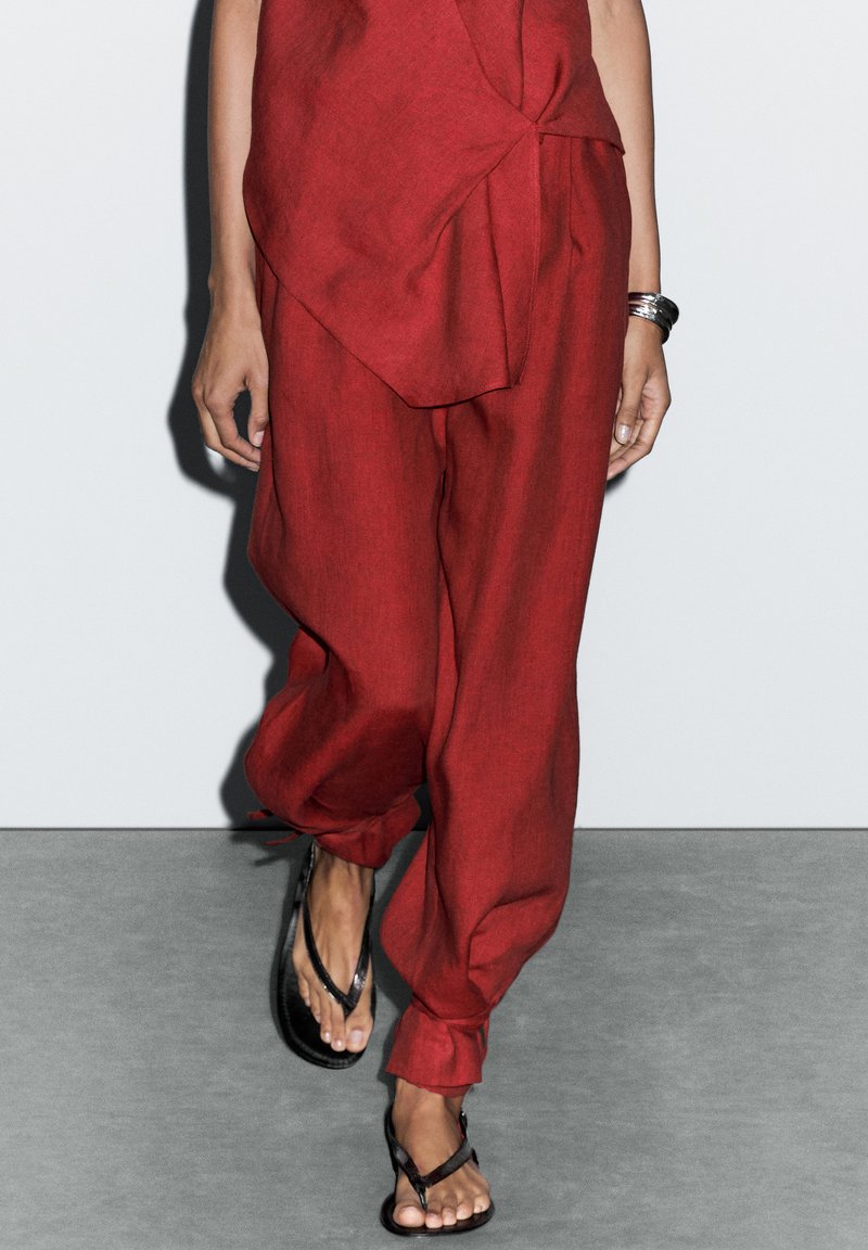 Pantaloni in lino rossi con una vestibilità ampia, con un dettaglio a nodo in vita. Abbinati a sandali neri. Sfondo grigio neutro.