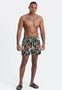 Homme avec tatouage d'aile sur la poitrine, portant un short de bain à motifs floraux et des sandales noires, debout devant un fond blanc uni.