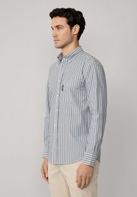 Camisa de botones a rayas en azul y blanco, con cuello, mangas largas y un bolsillo en el pecho. El material parece suave y tiene un diseño ajustado.