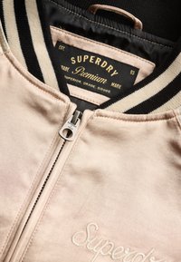 Beige Superdry-jas met zwart-witte gestreepte kraag, zichtbare ritssluiting, geborduurd logo op de borst en gebrandmerkt binnenlabel.