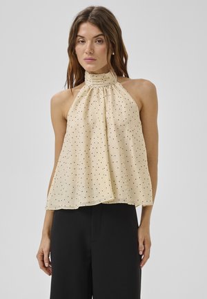 Frau trägt beige ärmelloses Top mit Neckholder und schwarzen Polka-Dots sowie schwarze Hochbundhose, steht vor schlichtem Hintergrund.