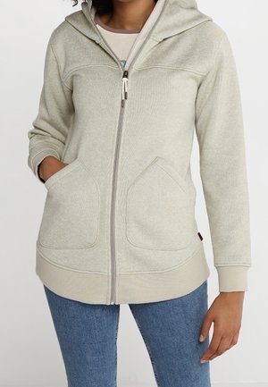 Frau trägt eine hellbeige Kapuzenjacke mit Reißverschluss und Vorderentaschen sowie blaue Jeans, steht mit einer Hand in der Tasche und der anderen seitlich am Körper.