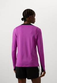 Lila hosszú ujjú sportfelső karcsúsított szabással és fekete nyakkivágással, fekete rövidnadrággal párosítva. Simaszövetű, sportoláshoz ideális.