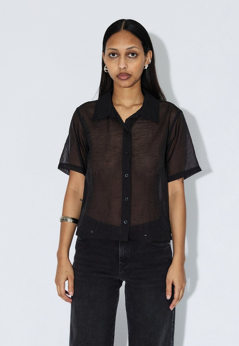 Camicia nera trasparente a maniche corte con colletto a punta, bottoni frontali e vestibilità rilassata, abbinata a pantaloni di denim scuro. Design semplice e strutturato.