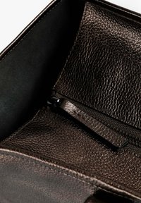 Interior de un bolso de cuero marrón con un acabado texturizado. Cuenta con un bolsillo negro con cremallera contra un forro suave. Sin adornos adicionales.
