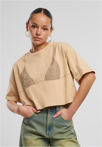 Lühikeste varrukatega lõikega beež t-särk, millel on minimaalistlik pruun graafika bikiinide topist. Valmistatud pehmest puuvillast, millel on mugav ja lõdvestunud istuvus.