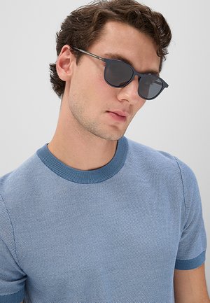 Giovane uomo con capelli castani corti che indossa occhiali da sole scuri e una camicia blu a maniche corte con trama, su uno sfondo chiaro e uniforme.