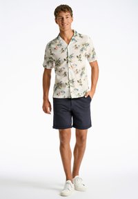 Camicia a maniche corte, di colore chiaro, con un motivo floreale e dettagli verdi e rosa, abbinata a pantaloni corti blu navy e sneakers bianche. Outfit estivo casual.
