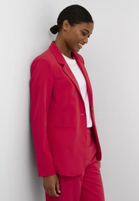 Blazer rosso con un design su misura, frontalmente con un bottone singolo, rever a punta e tasche laterali, abbinato a una t-shirt bianca e pantaloni coordinati.