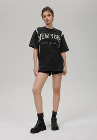 Svart oversized T-skjorte med teksten "NEW YORK", korte ermer og blonde detaljer på skuldrene. Brukt sammen med svarte shorts og ankelstøvletter.