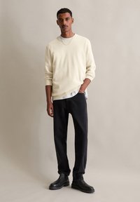Pull crème clair avec une texture en tricot, associé à un jean en denim sombre et des bottines noires. Le mannequin se tient avec confiance, les mains dans les poches.