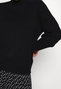 Schwarzer Rollkragenpullover mit strukturierten, gemusterten Ärmeln und geripptem Saum, kombiniert mit einem schwarz-weißen Polka-Dot-Rock.