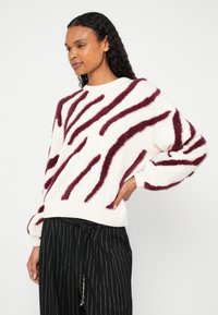 Fehér pulóver bolyhos bordó csíkokkal, laza szabással, kerek nyakkal és bordázott mandzsettákkal, fekete tűsarkú csíkos nadrággal párosítva.