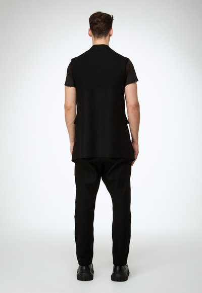 Schwarze ärmellose Weste über einem schwarzen T-Shirt, kombiniert mit schwarzen Hosen und klobigen schwarzen Stiefeln. Glatter Stoff mit minimalistischem Design.