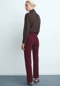 Femme aux cheveux blonds en chignon, portant un blouse à manches longues marron foncé, un pantalon taille haute bordeaux et des sandales à talons noires, se tenant de profil.