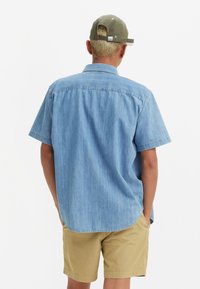 Blue denim short-sleeve shirt with a straight cut, visible stitching, and rounded hem; paired with beige shorts. Back view, cap on head.