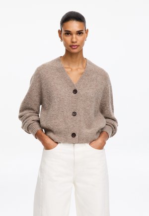 Frau mit zurückgebundenem dunklem Haar, die eine beige Strickjacke mit Knöpfen und weiße Hose trägt, steht mit den Händen in den Taschen vor weißem Hintergrund.
