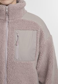 Približeni prikaz osobe u svijetlo ljubičastoj teksturiranoj flis jakni s džepom na prsima sa zatvaračem i podstavljenim ramenim panelima.