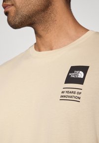 Μπεζ βαμβακερό T-shirt με στρογγυλή λαιμόκοψη, με μαύρο λογότυπο και κείμενο που αναγράφει «60 ΧΡΟΝΙΑ ΚΑΙΝΟΤΟΜΙΑΣ» εμφανώς.