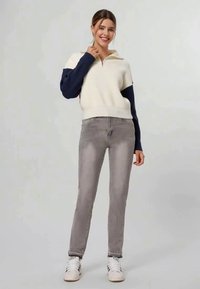 Pull zippé blanc et marine à texture côtelée, associé à un jean slim gris clair et des baskets blanches, posé devant un fond neutre.