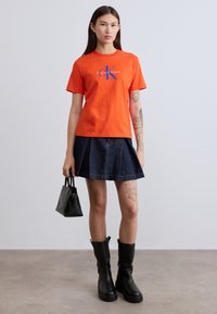 Πορτοκαλί βαμβακερό t-shirt με το λογότυπο "Calvin Klein" σε μπλε, συνδυασμένο με σκουρόχρωμη τζιν φούστα με πιέτες και μαύρες μπότες μέχρι το γόνατο.