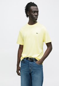 T-shirt di cotone giallo a maniche corte, con un piccolo logo verde sul petto a sinistra. Abbinata a jeans blu e una cintura nera.
