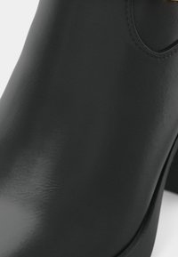 Crne kožne gležnjače s glatkom završnom obradom, zaobljenim vrhom i laganom petom. Sadrže minimalističke dizajnerske elemente i detalje šivanja.