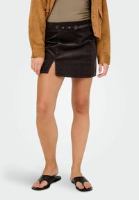 Donna che indossa una giacca in camoscio marrone, un top verde oliva, una mini gonna in velluto a coste nera con cintura e spacco, e sandali neri con punta aperta.