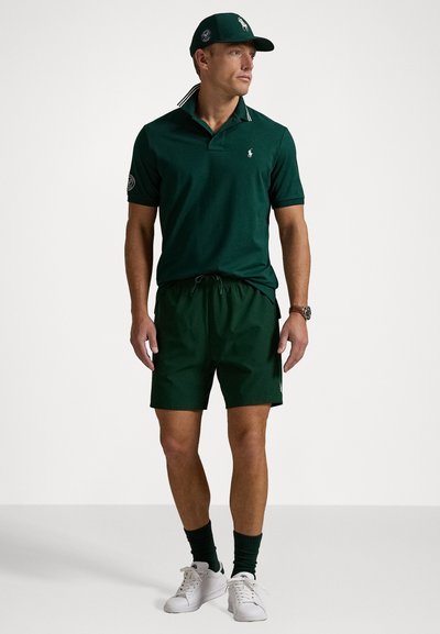 Polo verde cu guler în dungi, pantaloni scurți verzi asortați, șosete verzi și adidași albi. Căciula de baseball are un logo.