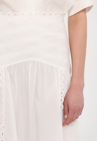 Robe en coton blanc avec un corsage texturé orné d'un bord en dentelle festonné et un jupon plissé froncé, mettant en valeur des détails délicats du tissu.