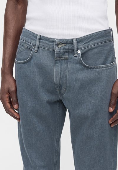 Personne portant un jean bleu en denim et un débardeur blanc côtelé, avec un focus sur la taille et les mains détendues sur les côtés.
