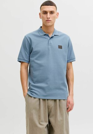 Jauns vīrietis stāv ar rokām kabatās, valkājot gaiši zilu īsroku polo krekliņu un bēšas vaļīgas bikses uz vienkrāsaina fona.