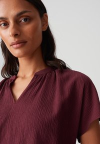 Blusa bordeaux a trama con scollatura a V, maniche corte e tessuto morbido e stropicciato. I dettagli includono tessuto arruffato sulle spalle.