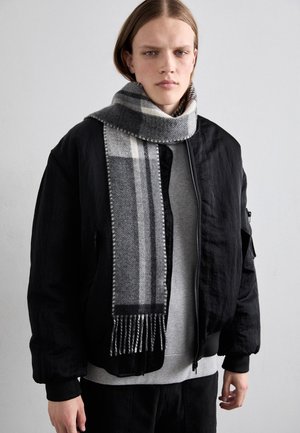 Jeune personne aux cheveux raides mi-longs portant une veste bomber noire, un pull gris et une écharpe à carreaux grise, devant un fond uni.