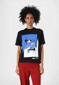 Melnas kokvilnas t-krekls ar zilu un baltu grafisko attēlu, kurā redzama figūra brillēs, apakšā atrodas "KARL LAGERFELD JEANS" logo.