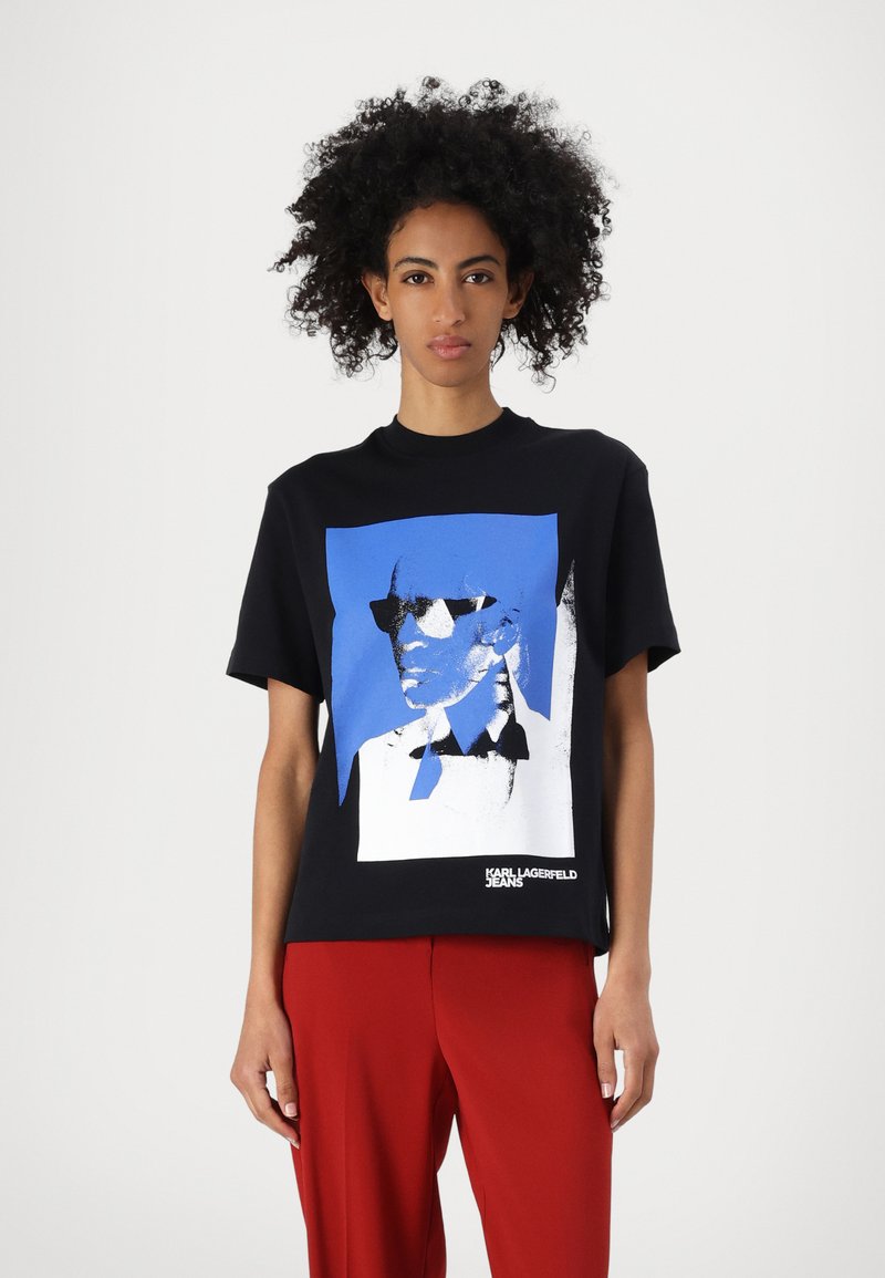 Melnas kokvilnas t-krekls ar zilu un baltu grafisko attēlu, kurā redzama figūra brillēs, apakšā atrodas "KARL LAGERFELD JEANS" logo.