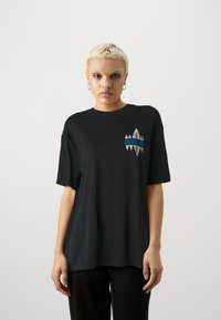 Mustat ylisuurent t-paita, joka on valmistettu sileästä kankaasta. Paidassa on monivärinen logo, jossa on geometrisia elementtejä vasemmalla rintapielessä.