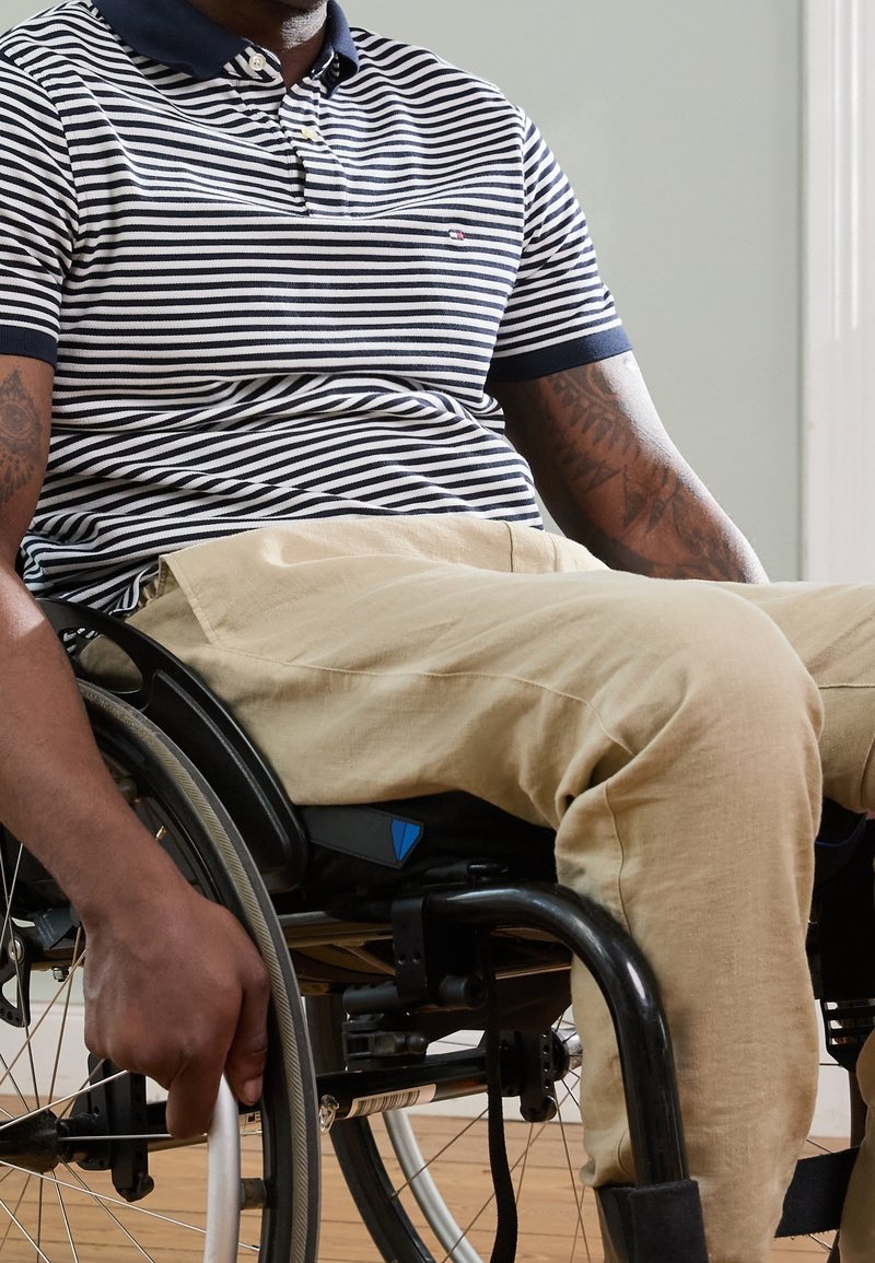 Homme en chemise rayée et pantalon beige utilisant un fauteuil roulant manuel, tenant la roue dans une pièce aux murs clairs et au sol en bois.