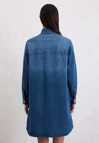 Denimhemdkleid in Mittelblau, mit langen Ärmeln, geknöpften Manschetten und einem geschwungenen Saum. Der Stoff ist glatt und hat ein klassisches Kragen-Design.