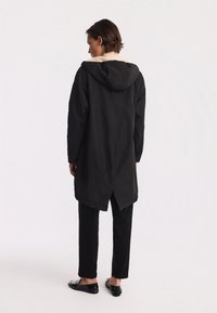 Czarna parka z kapturem i kremową podszewką, z dłuższym tylnym dołem i luźnym krojem. Połączona z czarnymi spodniami i butami.