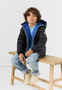 Giacca a vento nera con fodera blu, cappuccio e zip; abbinata a una maglietta polo azzurro chiaro, jeans in denim chiaro e sneaker beige.