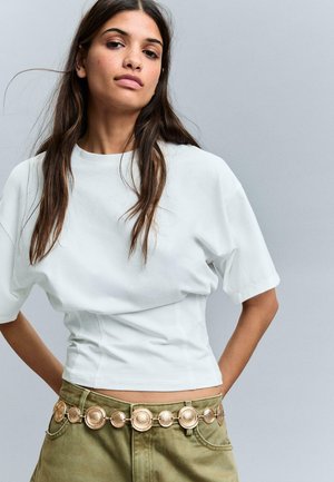 Hosszú barna haját viselő nő, fehér, rövidített pólót és olívazöld nadrágot visel, amelyet arany pénzérme öv díszít, szürke háttér előtt áll.