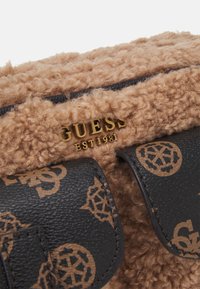 Hnědá texturovaná látková taška s černými koženými kapsami s potiskem a zlatým logem „GUESS EST 1981“.