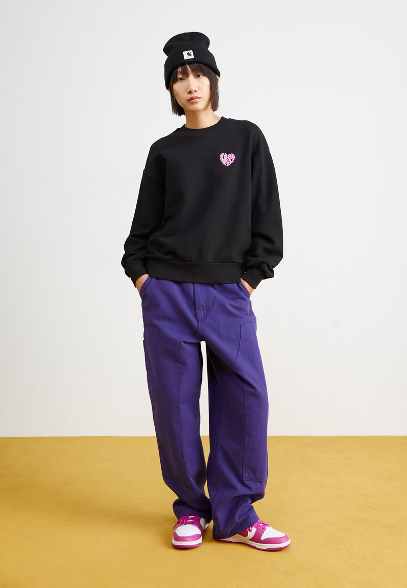 Sweatshirt noir avec un graphisme de cœur rose, associé à un pantalon cargo violet large et des baskets roses. Le modèle porte un bonnet noir.