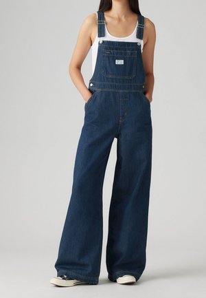 Salopette en denim bleu foncé avec jambes larges, bretelles réglables, poche avant et poches latérales. Associée à un débardeur blanc et des baskets.