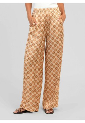 Pantalon beige à jambes larges avec motif floral rouge et jaune, porté avec des sandales noires, mains dans les poches et bord supérieur blanc visible.