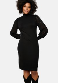Vestito nero a maglia con colletto alto e silhouette aderente. Presenta maniche lunghe in pizzo trasparente e decorazioni a coste sull'orlo.