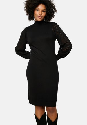 Vestido de punto negro con cuello alto y silueta ajustada. Presenta mangas largas de encaje transparente y estampado, además de detalles de ribete en el dobladillo.