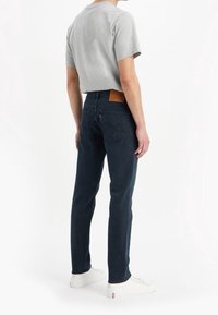 Mörkblå denimjeans med rak passform, med en beige läderlapp på bakre midjan och två bakfickor, tillsammans med en grå t-shirt.